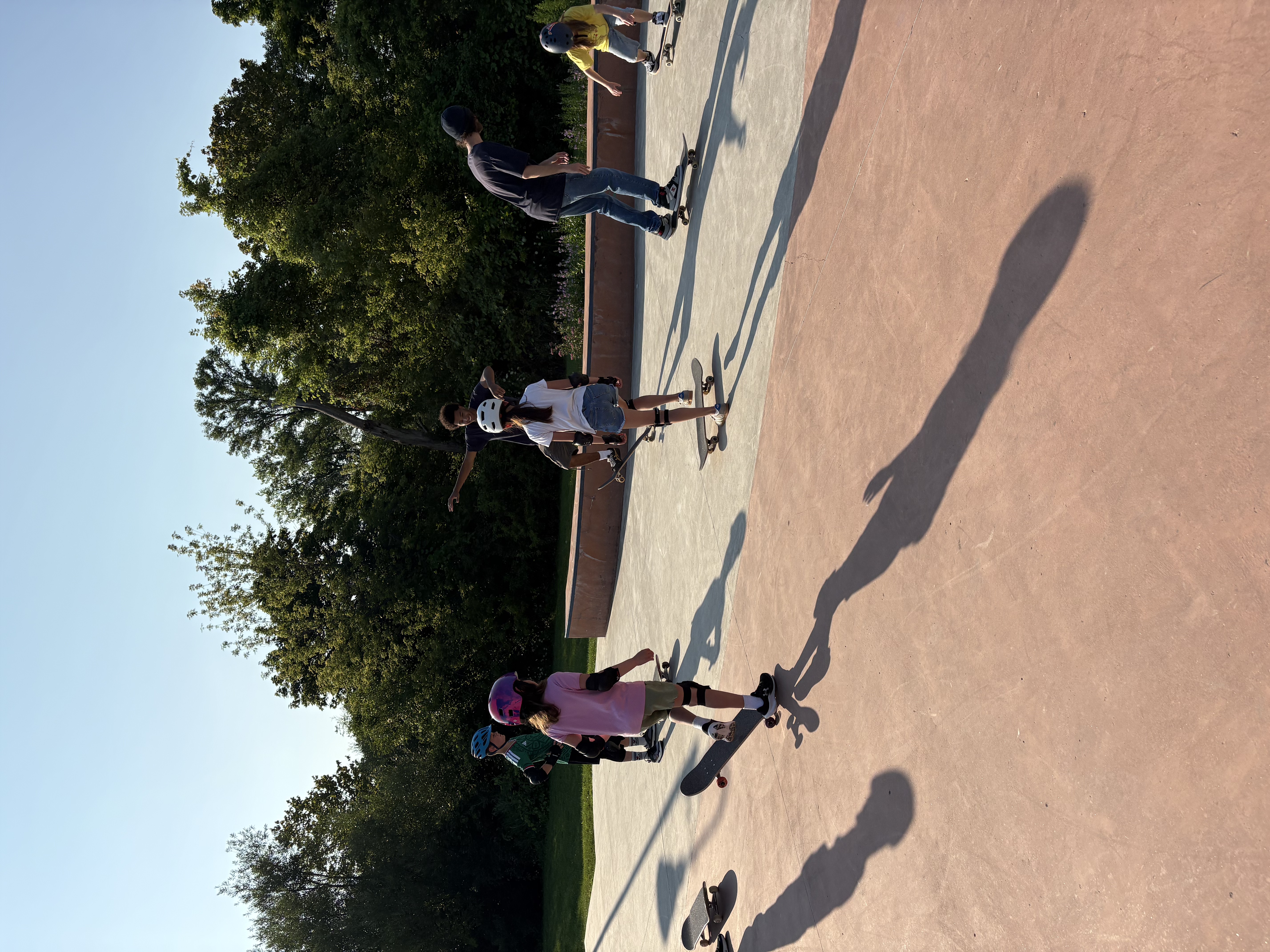 Kids and teens with skateboards standing on a concrete skate plaza at sunset, with a coach nearby