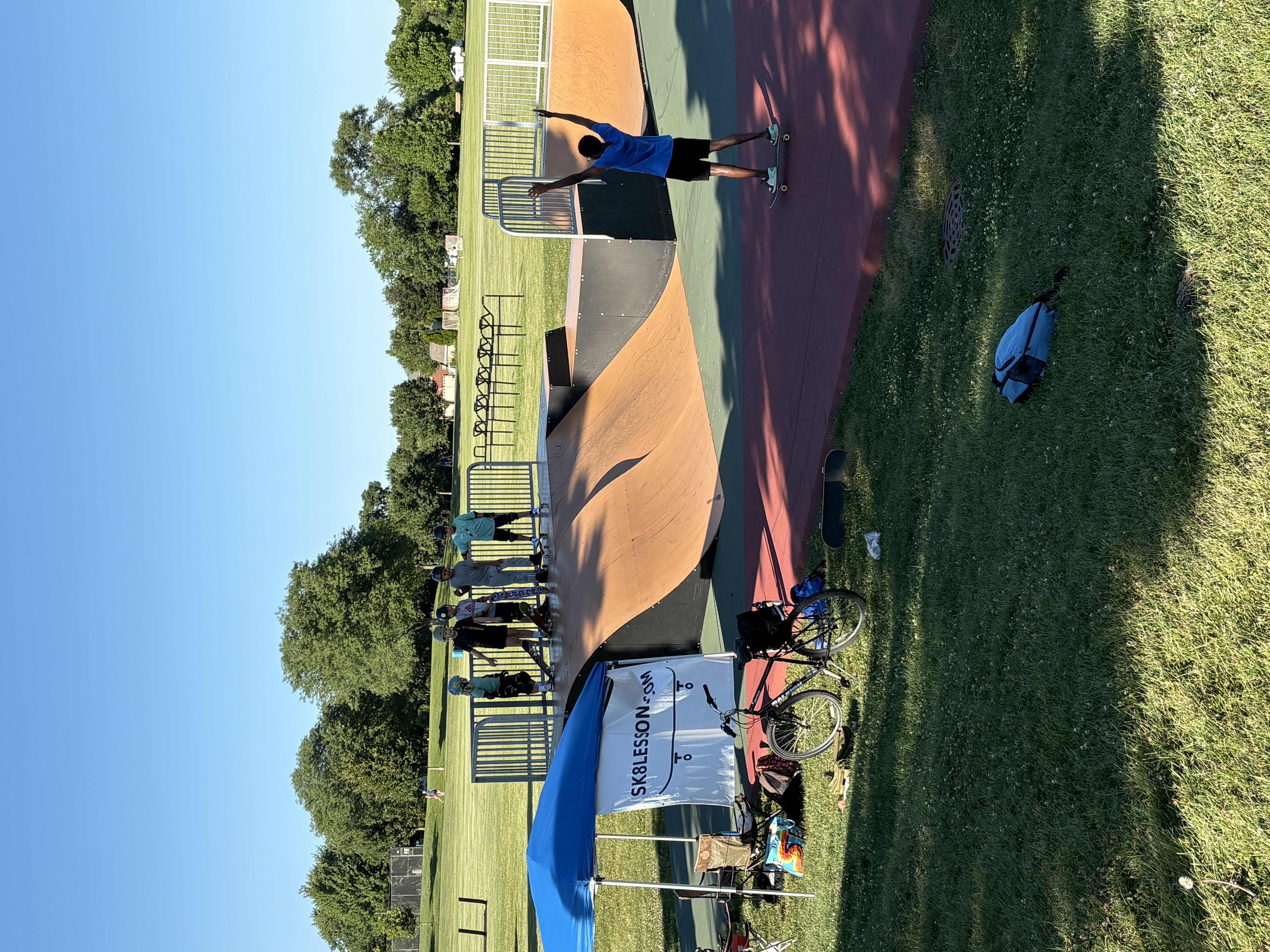 A skater standing at the top of a ramp with a coach and other young skaters watching, near a Sk8lesson.com tent at Newton Park