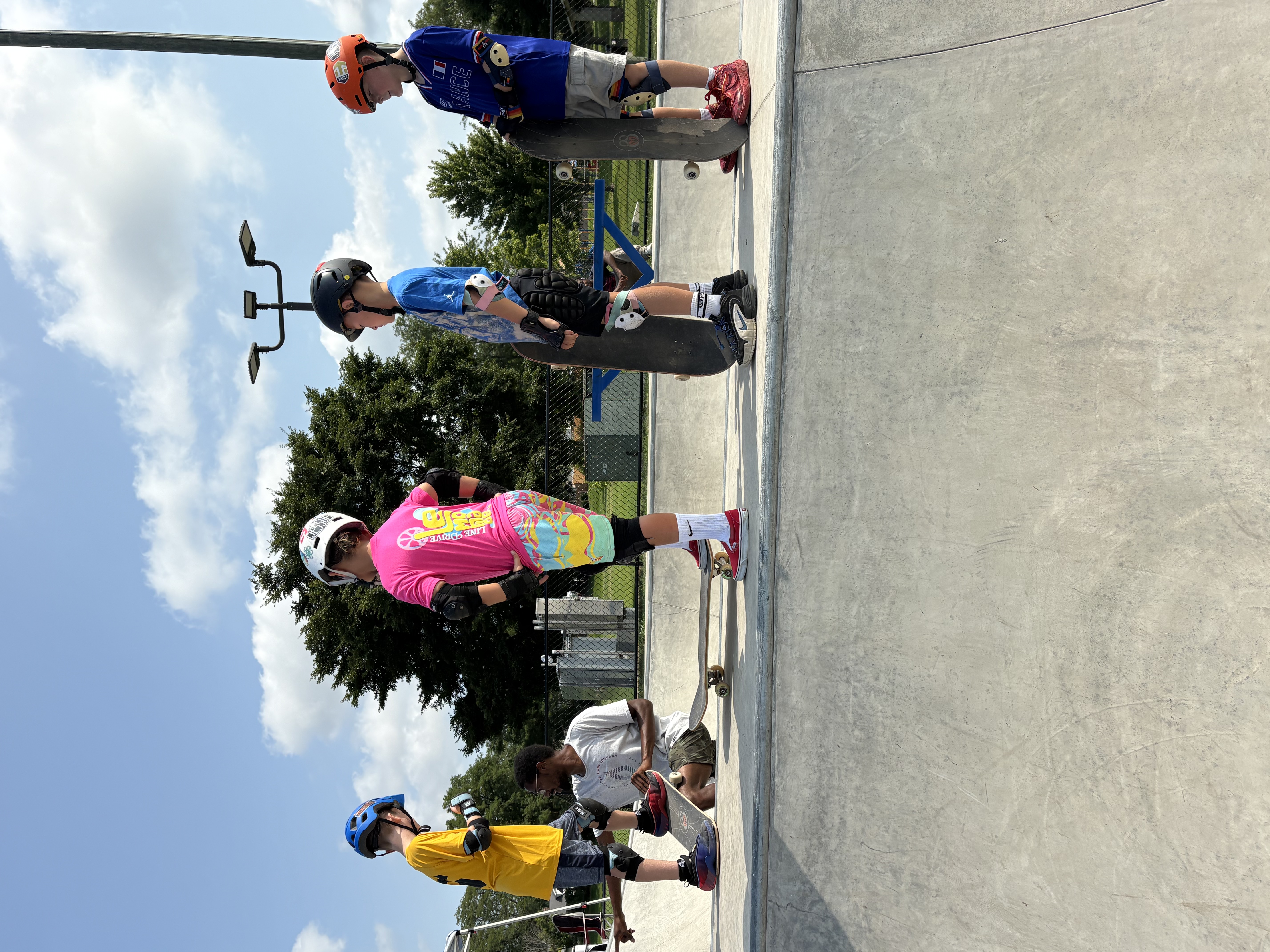 Four young skaters in helmets and pads standing at the edge of a skatepark bowl with their skateboards